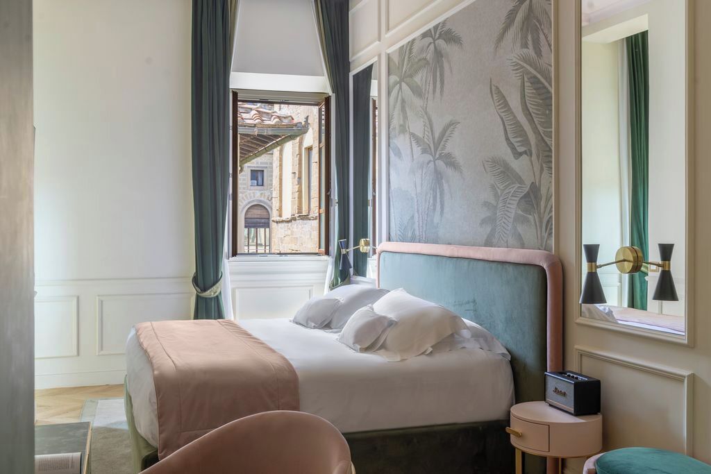 A bedroom with a bed, a nightstand, a chair, a window with curtains, and a view of a building outside.
