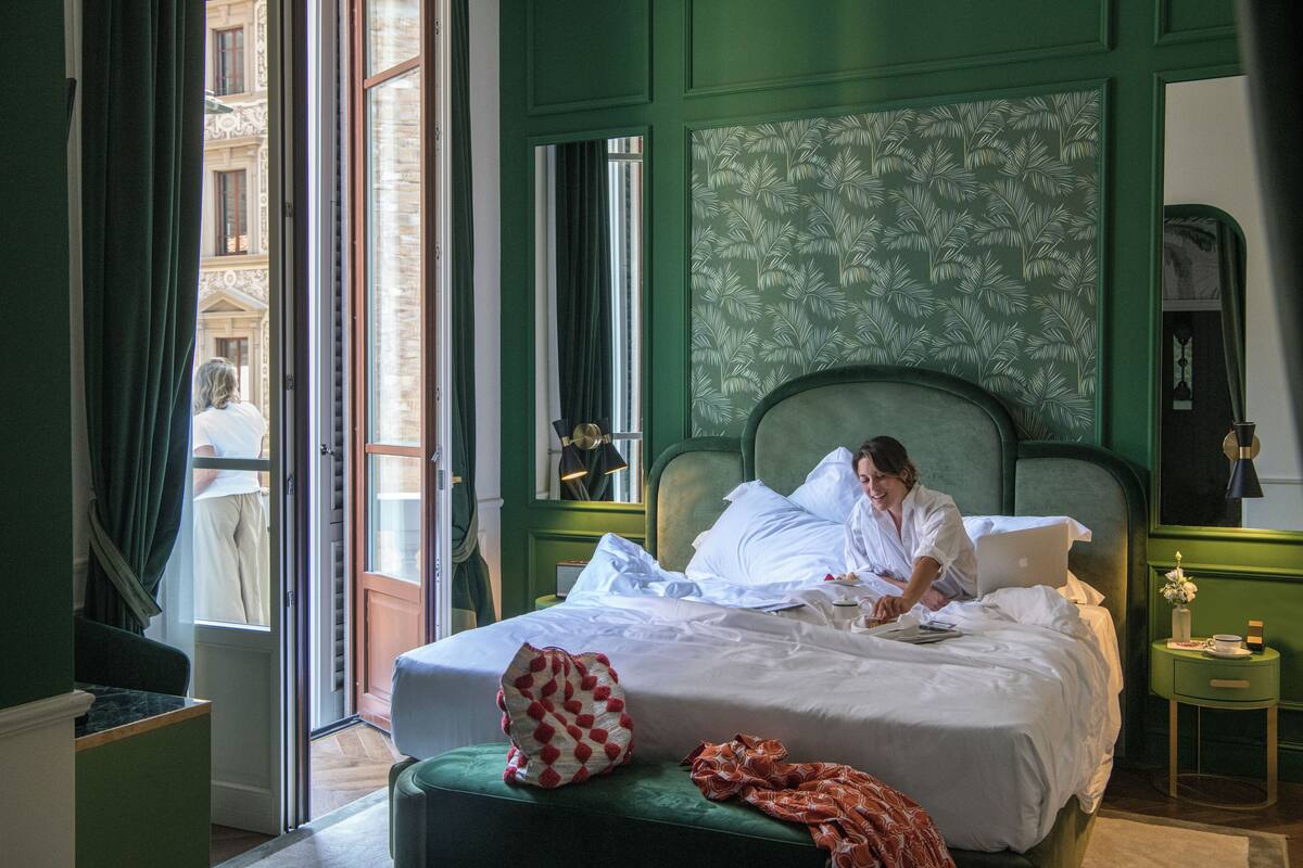 A hotel room with a large bed, a green upholstered bench, a small bedside table with a lamp, and a view of the street through an open window.
