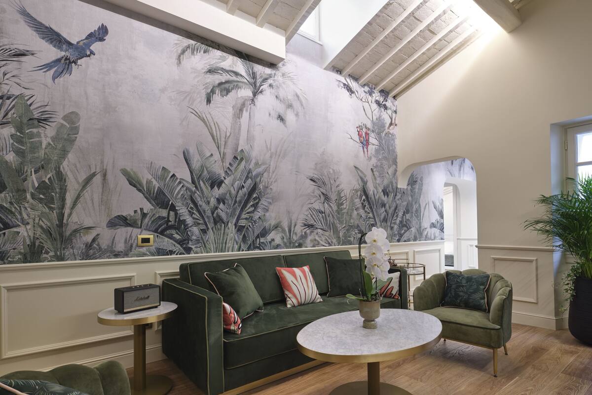 A modern living room with a green sofa, a round coffee table, and a green armchair. A large mural with tropical foliage and birds is on the wall.