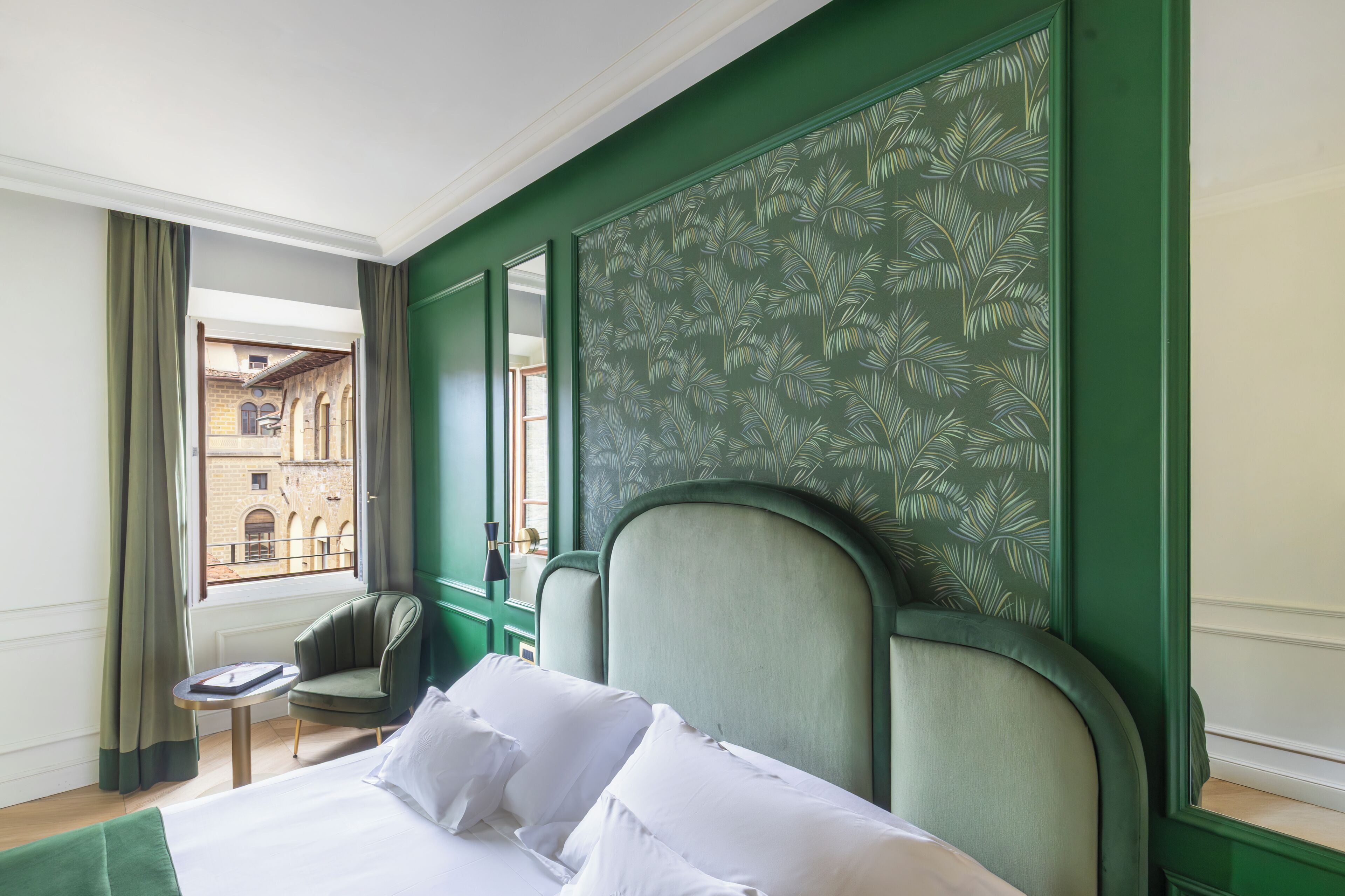 A hotel room with a bed, a chair, a small table, and a view of a building through a window.