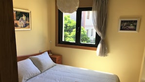 Una cama bien hecha con una colcha estampada, una mesita de noche con una lámpara y una ventana que da a vistas de vegetación.