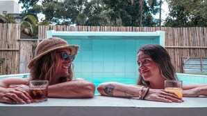 Two people sitting by a pool with drinks, surrounded by a bamboo fence and trees. - Indonesia Backpacker Hostels  (Kuta)