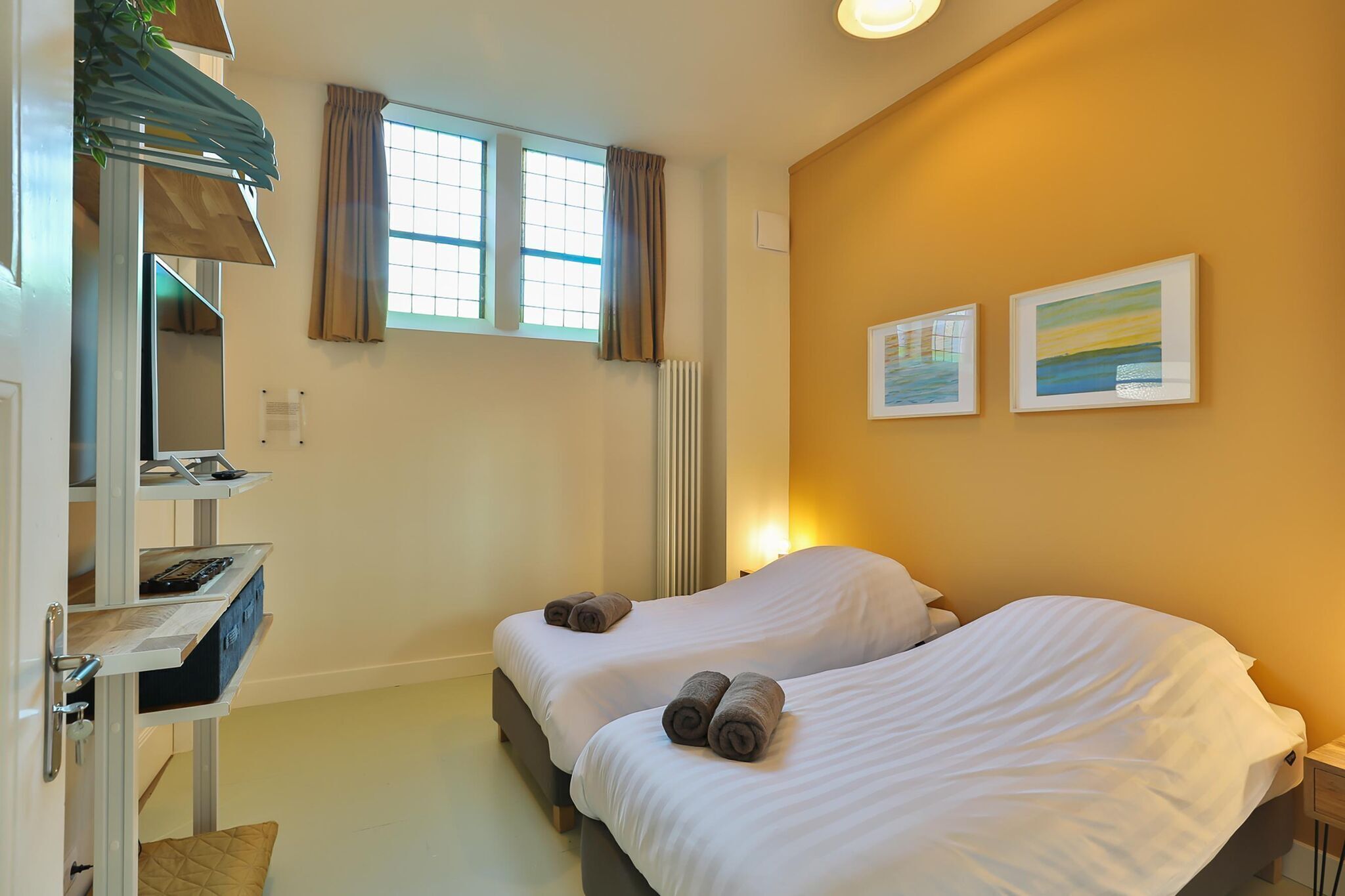 A hotel room with two beds, a desk with a computer, a radiator, and two framed pictures on the wall.