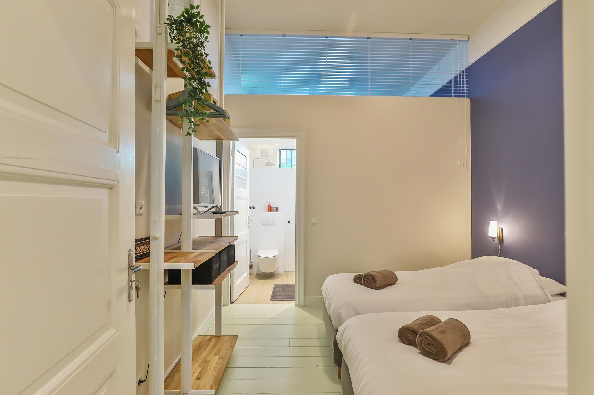 Une chambre d’hôtel avec deux lits, un bureau avec un ordinateur, une fenêtre avec des stores et une salle de bain visible à travers une porte ouverte.