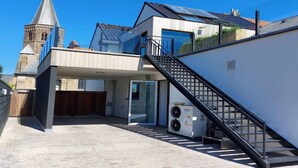 Une maison moderne avec un escalier menant à un espace extérieur couvert, un grand climatiseur et, au loin, le clocher d’une église.