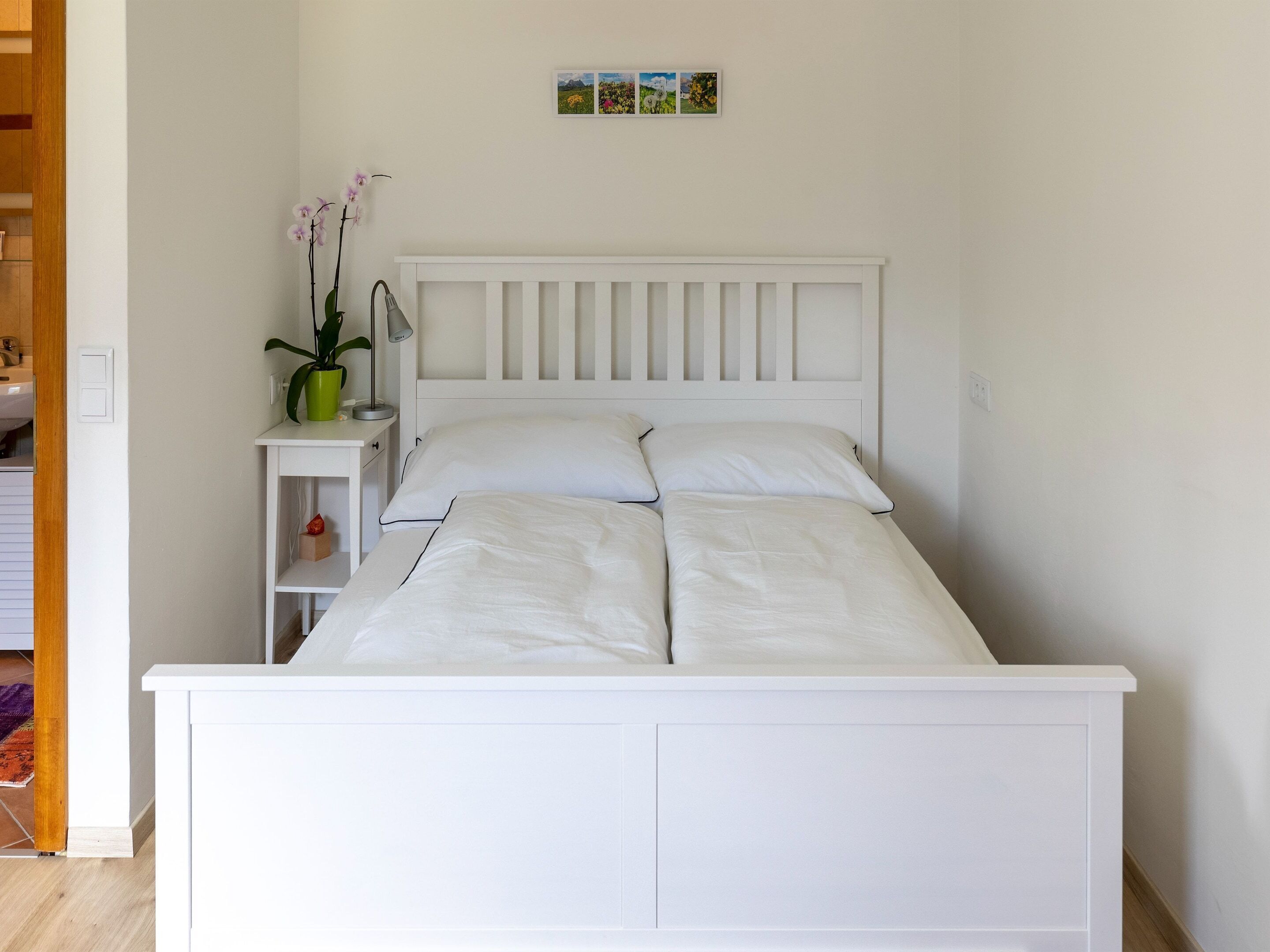 A white bed with a headboard, a bedside table with a lamp, and a small shelf with a plant.