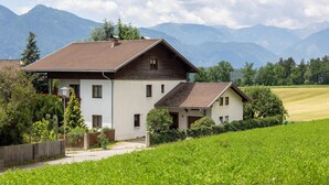 Una casa de dos pisos con techo inclinado, rodeada de abundante vegetación y montañas al fondo.