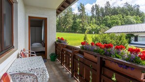 Un balcón con mesa y sillas, baranda de madera y vista a un campo verde y árboles.