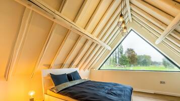 Un dormitorio con techo inclinado, una cama con ropa de cama oscura, una lámpara de noche y vistas al exterior con vegetación.