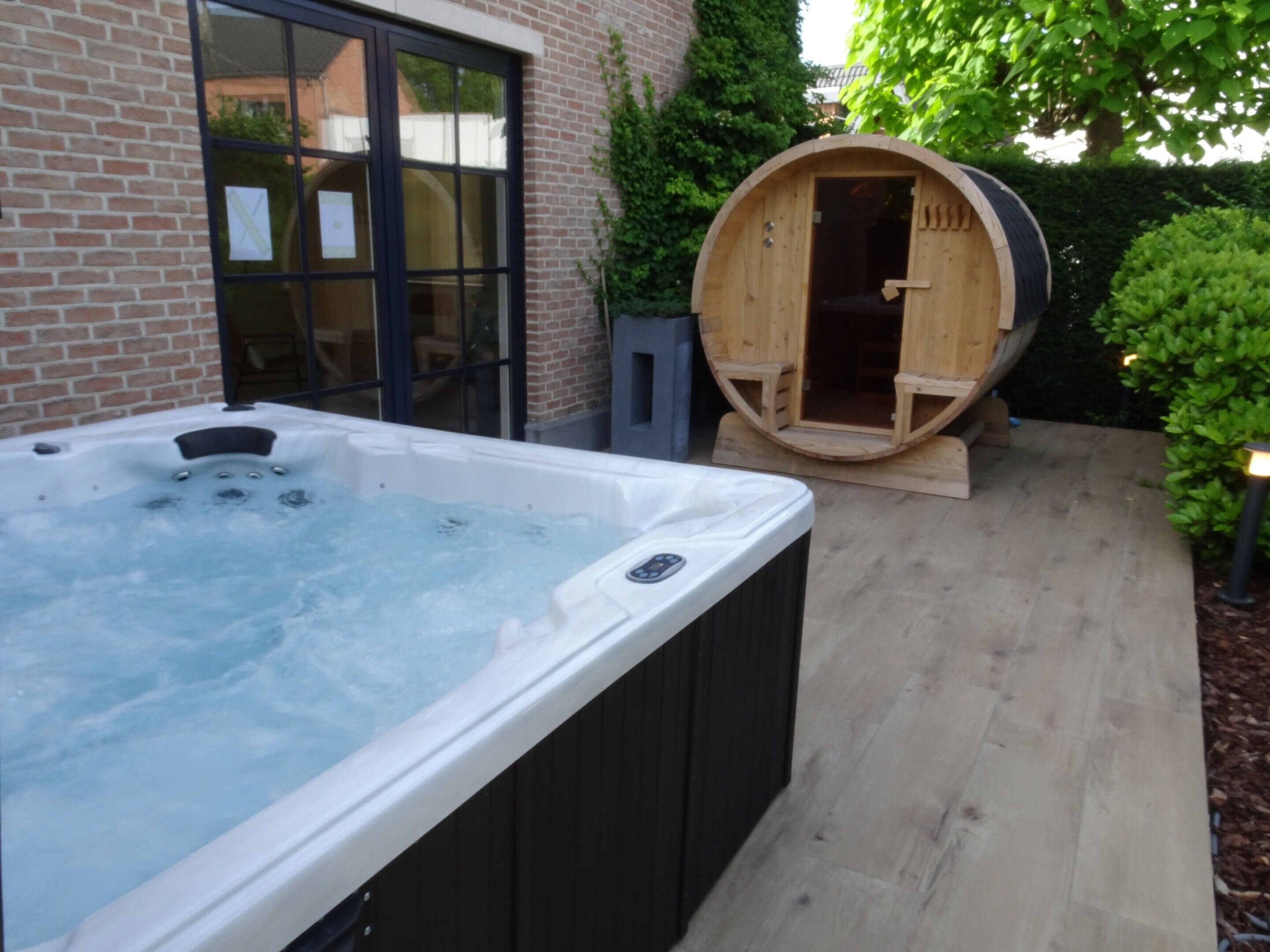 A hot tub and a wooden barrel sauna in a backyard with a brick wall and potted plants.