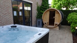 Un jacuzzi y una sauna de barril de madera en un patio trasero con un muro de ladrillo y plantas en macetas.