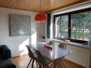 Un espace repas comprenant une table, des chaises, un luminaire suspendu, une fenêtre donnant sur des arbres et un mur orné d’un motif géométrique.