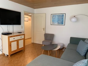 Un salon avec un téléviseur à écran plat, un canapé gris, une petite table, un fauteuil gris et un tableau accroché au mur.