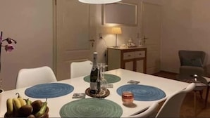 A dining table with a white tablecloth, blue and green circular placemats, and a coffee machine. There is a lamp on the wall, a chair, and a small cabinet with a lamp on it. - Holiday Apartment in Beautiful Hunningen (Buellingen)