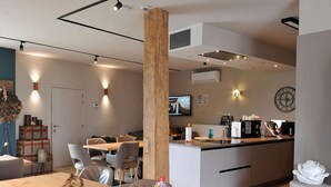 A modern dining area with a wooden table, chairs, and a central pillar. A television is mounted on the wall, and a clock is visible. - Vlaanderen Cozy Cottage (Maasmechelen)