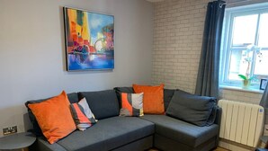 Ein modernes Wohnzimmer mit einer grauen Eckcouch, orangefarbenen und grauen Kissen, einem kleinen runden Tisch und einem farbenfrohen abstrakten Gemälde an der Wand.
