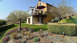 A two-story house with a balcony, a wooden facade, and a well-maintained garden with hedges and flowering plants. - Holiday Home in Noiseux With BBQ Terrace (Somme-Leuze)