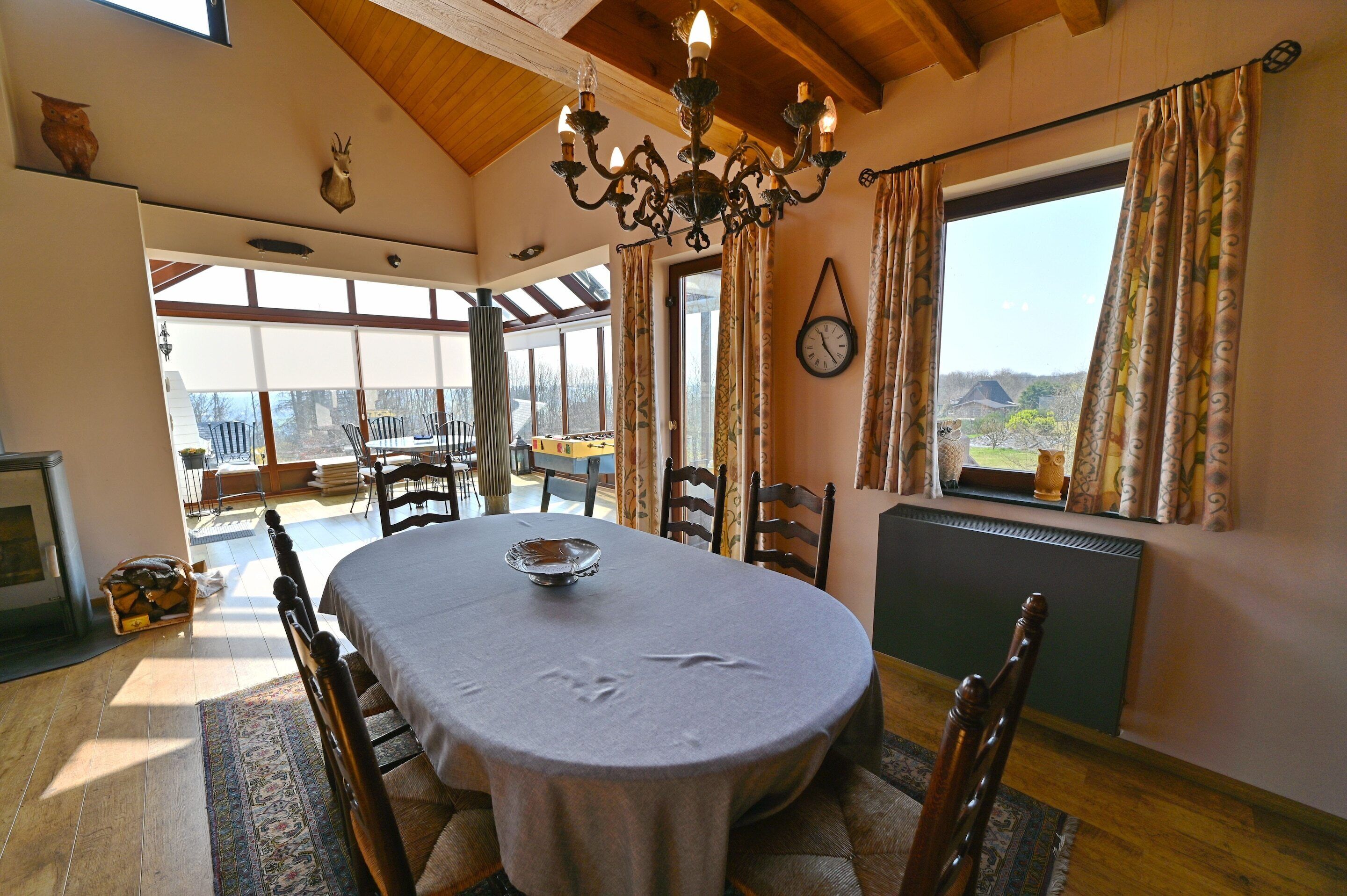 A dining room with a round table, chairs, a chandelier, and a view through large windows.