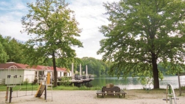 A sandy beach area with playground equipment, a picnic table, and a lake in the background. - Welcoming Chalet in Rekem Tucked in Nature (Lanaken)