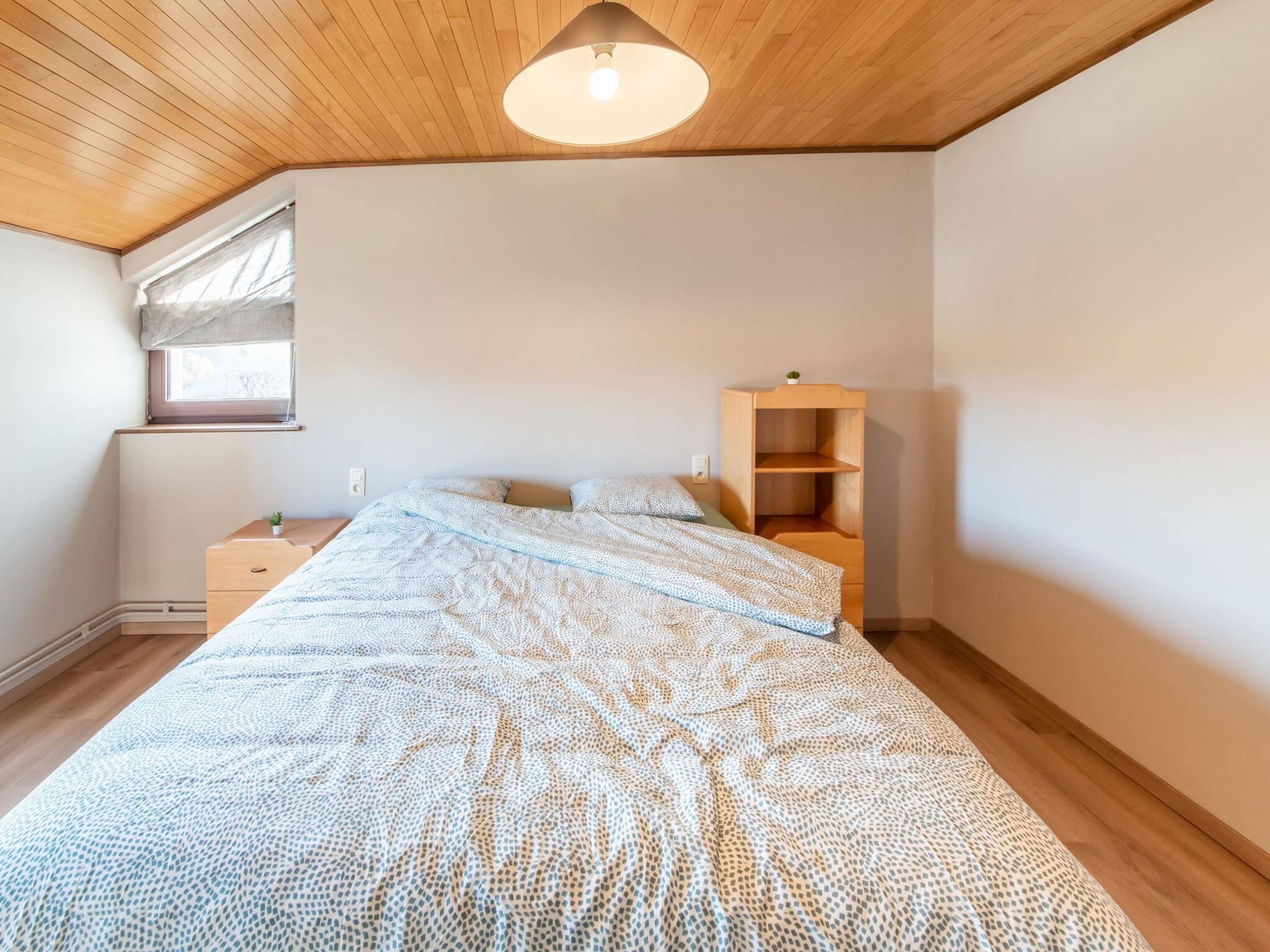 A bedroom with a wooden ceiling, a bed with a patterned bedspread, a nightstand, a small wooden shelf, and a window with a curtain.