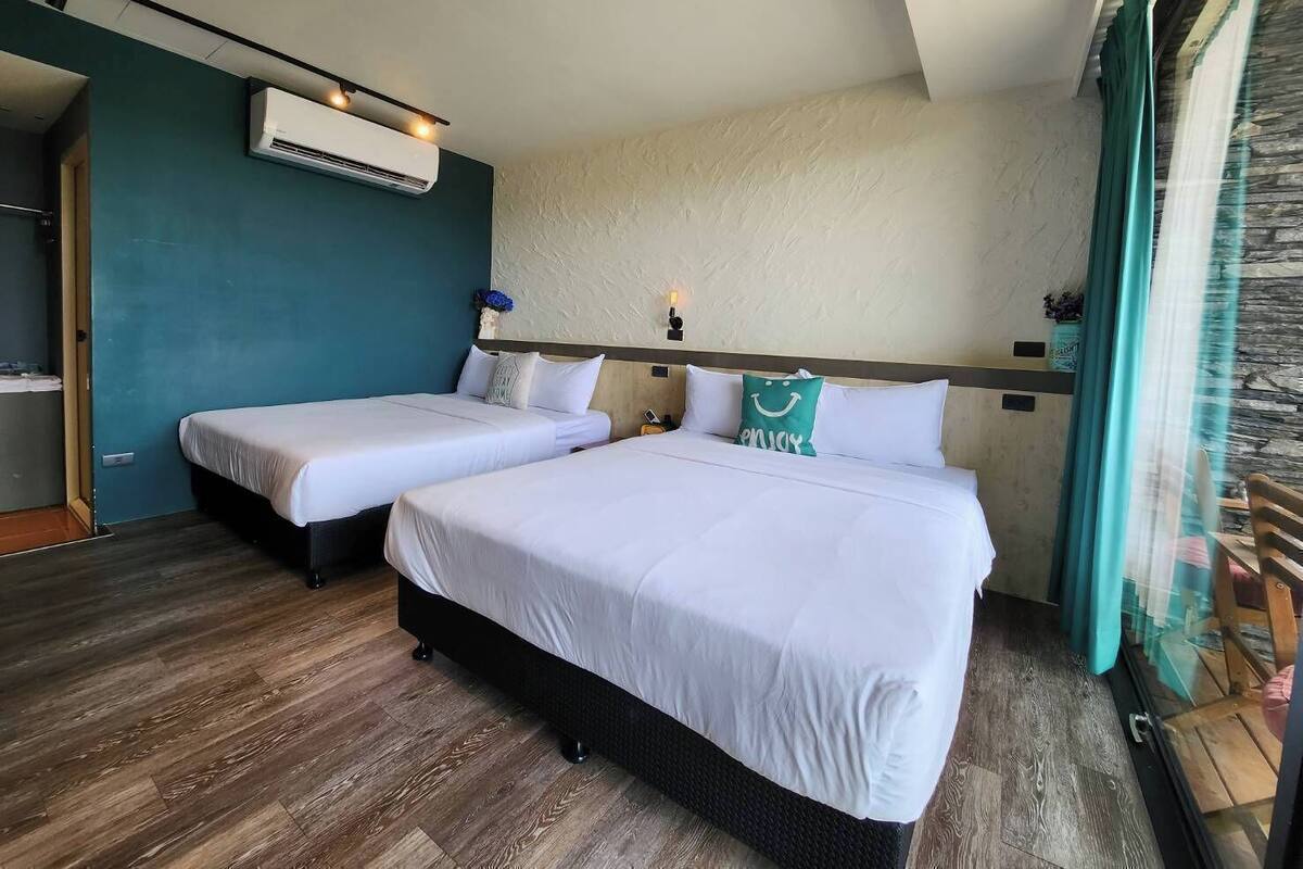 A hotel room with two beds, an air conditioning unit, a wall-mounted light, and a view of the outside through a window with curtains.