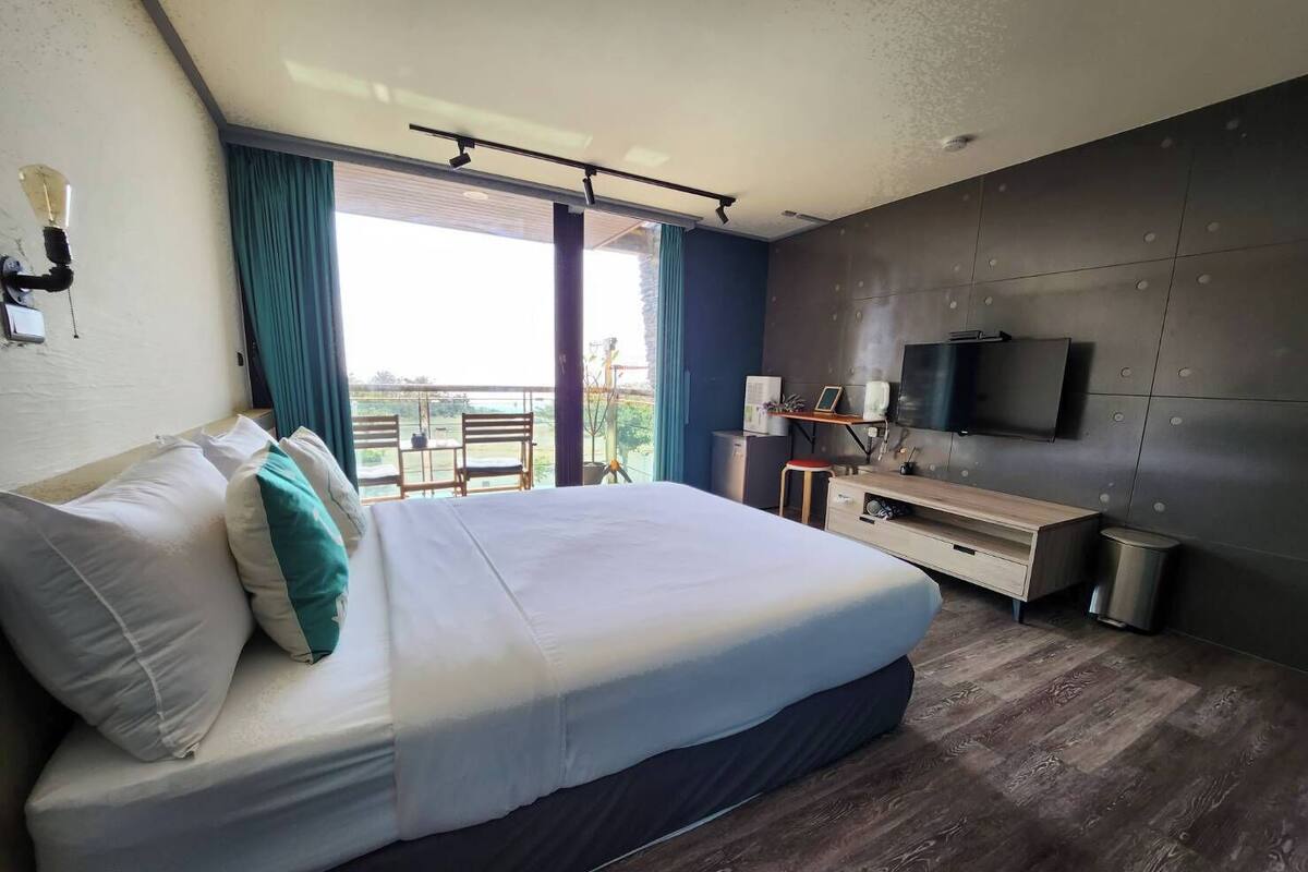 A modern hotel room with a large bed, a desk, a TV, and a balcony with a view.