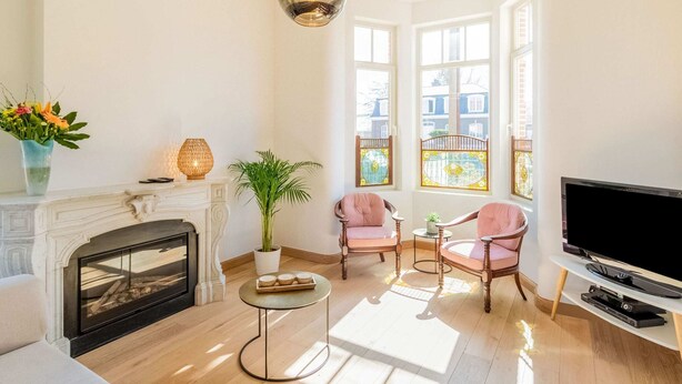 Un salon doté d’une cheminée, de deux fauteuils roses, d’une table basse ronde, d’un téléviseur et de grandes fenêtres offrant une vue sur la ville.
