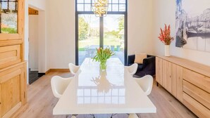 Un espace repas moderne avec une table et des chaises blanches, un meuble en bois, un canapé noir et une grande fenêtre donnant sur un paysage verdoyant.