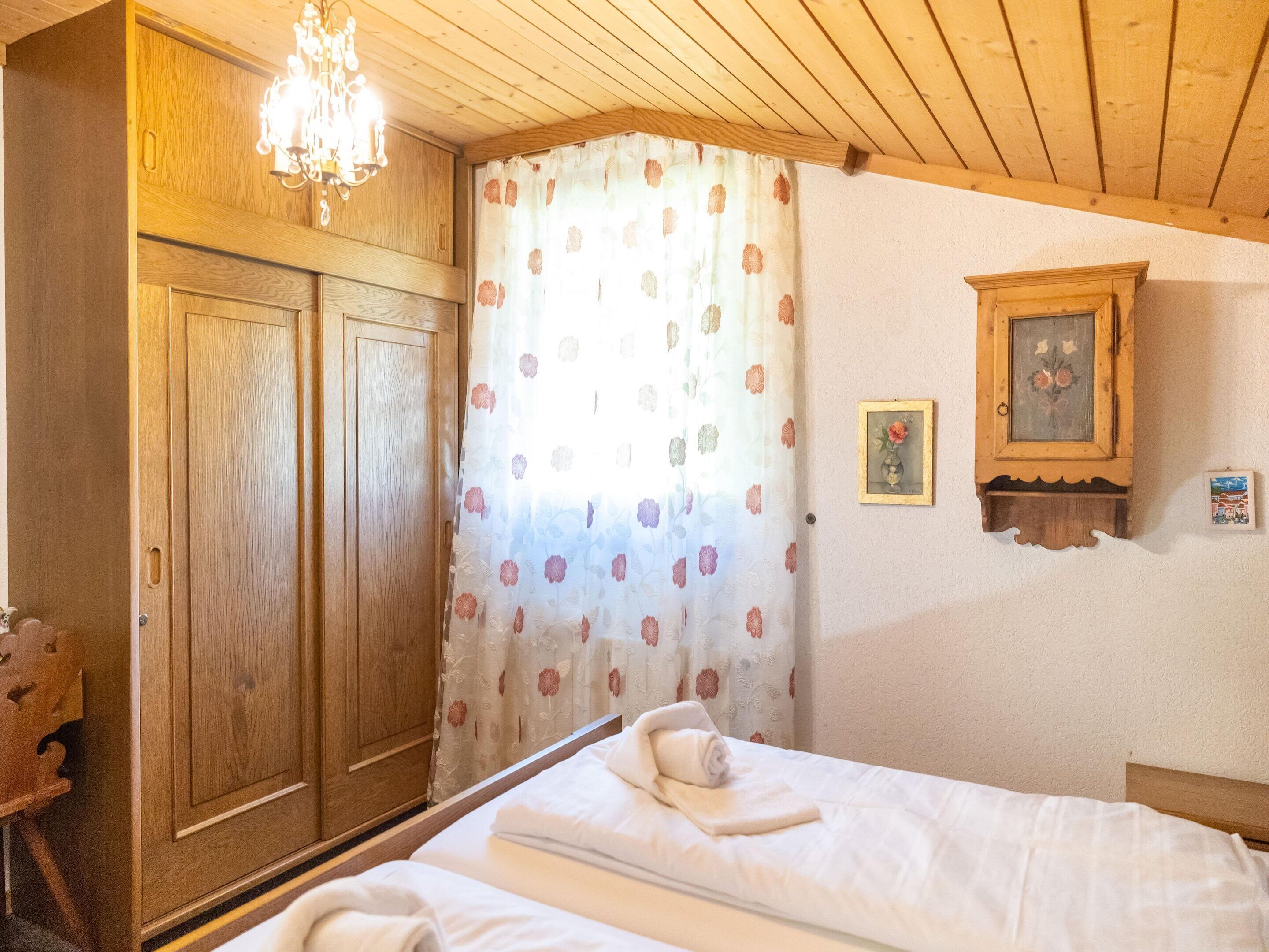 A bedroom with wooden furniture, a bed with white linens, a wooden cabinet, a chandelier, and a floral curtain.