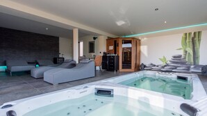 A modern spa area with two hot tubs, a gray sectional sofa, and a wooden sauna. - Holiday Home in Vlijtingen With Sauna (Riemst)