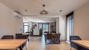 A modern dining area with a long wooden table and chairs, a reception desk, and a large window with blinds. - Holiday Home in Vlijtingen With Sauna (Riemst)