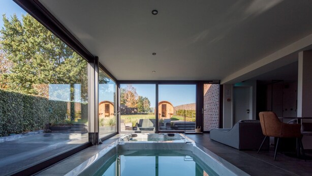 A modern indoor swimming pool with glass walls, a seating area, and a view of the surrounding landscape. - Holiday Home in Vlijtingen With Sauna (Riemst)