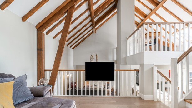 A spacious loft with a high ceiling, exposed wooden beams, a grey sofa, and a flat-screen TV mounted on the wall. - Alken Farm Stay With Terrace (Alken)
