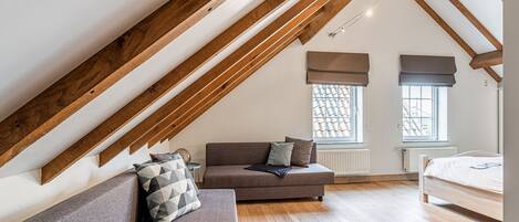 A loft bedroom with a sloped ceiling, wooden beams, a sofa, a bed, and two windows with blinds.