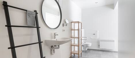 Un baño moderno con un lavamanos blanco, un espejo redondo, un perchero negro y una estantería de madera.