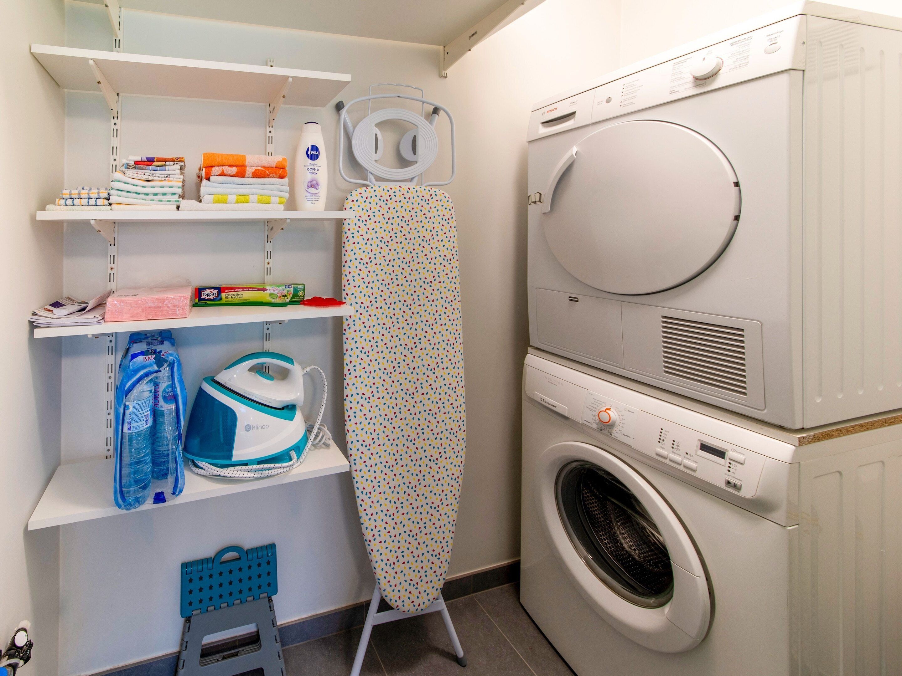 A laundry area with a washing machine, ironing board, shelves with folded clothes, and ironing supplies.