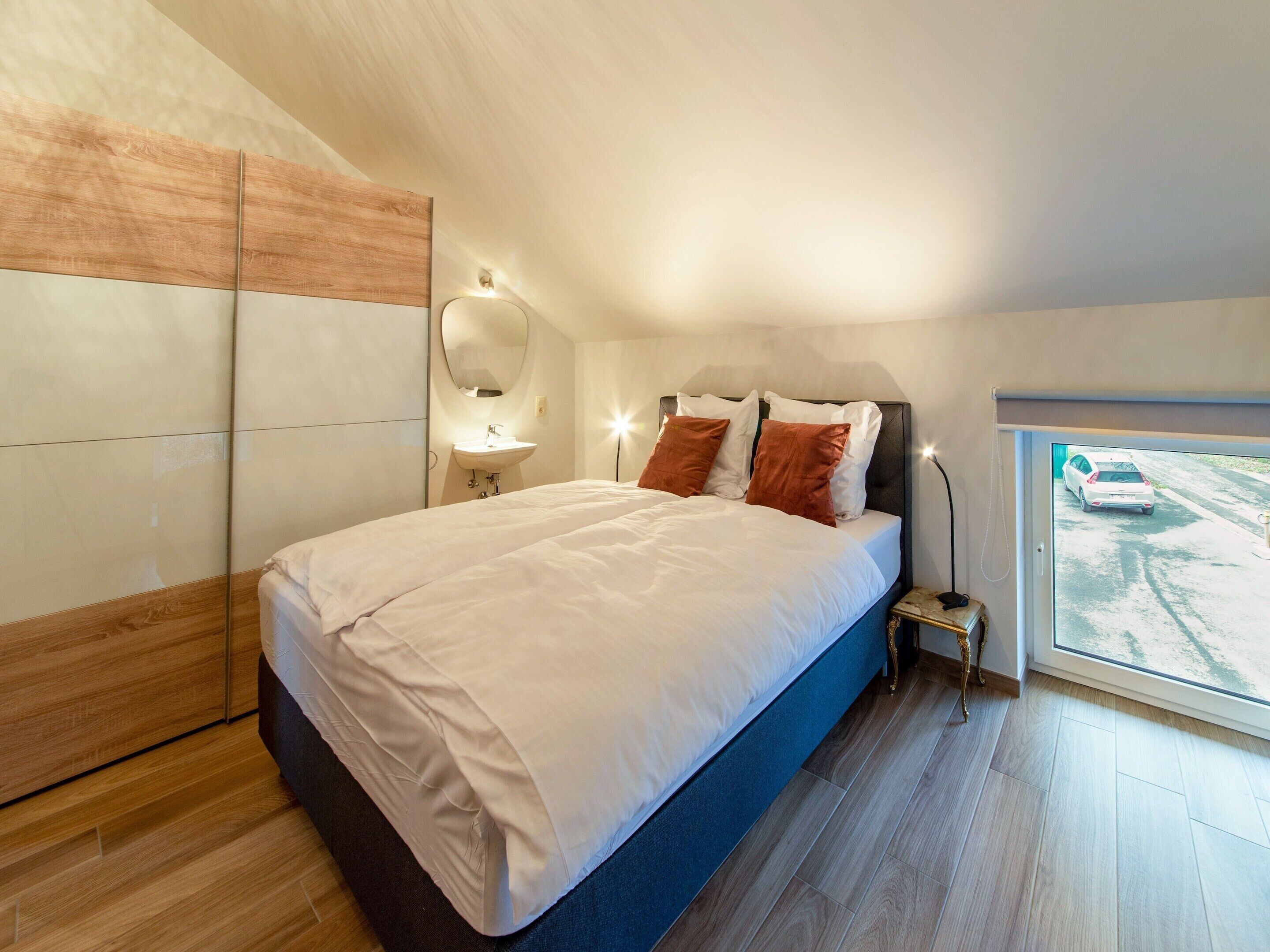 A modern bedroom with a large bed, a bedside table, a mirror, and a window with a view of a car.
