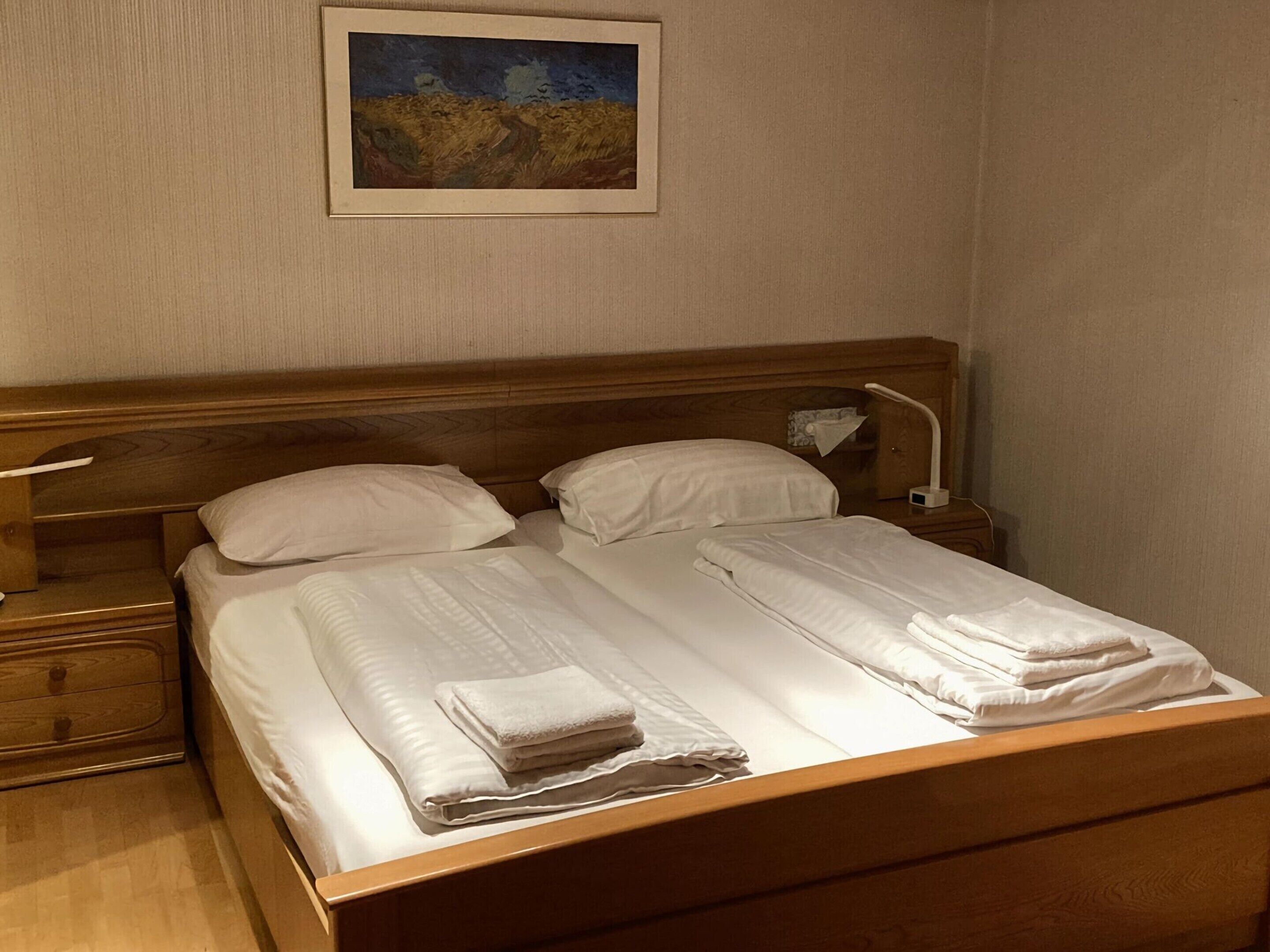 Una cama bien hecha con sábanas blancas y toallas dobladas, cabecera de madera, mesita de noche con lámpara y un cuadro en la pared.