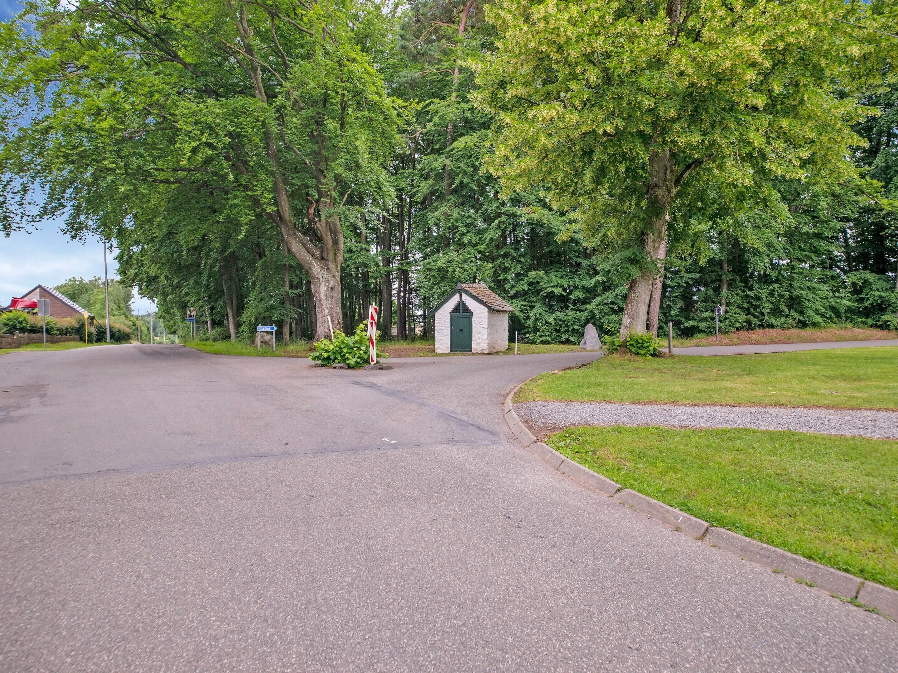 A rural road with a small stone building, surrounded by lush green trees and a grassy area.