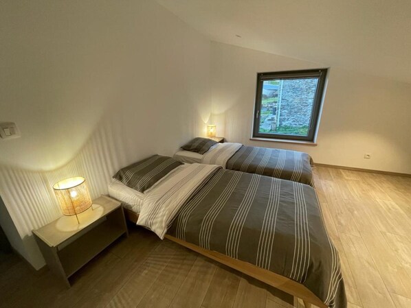 A bedroom with two beds, a bedside table with a lamp, and a window with a view of a stone wall. - Holiday Home in Walloon Ardennes (Bievre)
