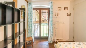 Ein Zimmer mit Holzboden, einem Bett mit gemustertem Überwurf, einem Sessel, einem Bücherregal, einem Heizkörper und einer Tür zu einem Balkon mit Blick auf ein Gebäude.