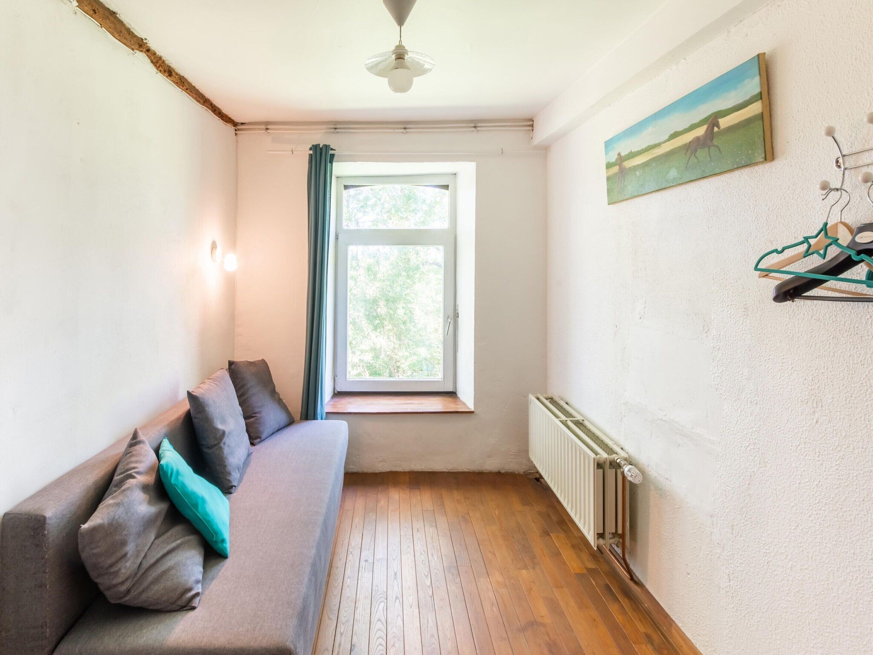 A room with a wooden floor, a grey sofa with cushions, a window with curtains, a radiator, and a painting on the wall.