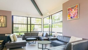 Ein modernes Wohnzimmer mit einem Ecksofa, einem runden Couchtisch und großen Fenstern mit Blick auf Grünflächen.