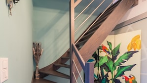 Un escalier avec des marches en bois et des rampes en métal, menant à une pièce ornée d’une fresque représentant des perroquets aux couleurs vives et des feuilles tropicales.