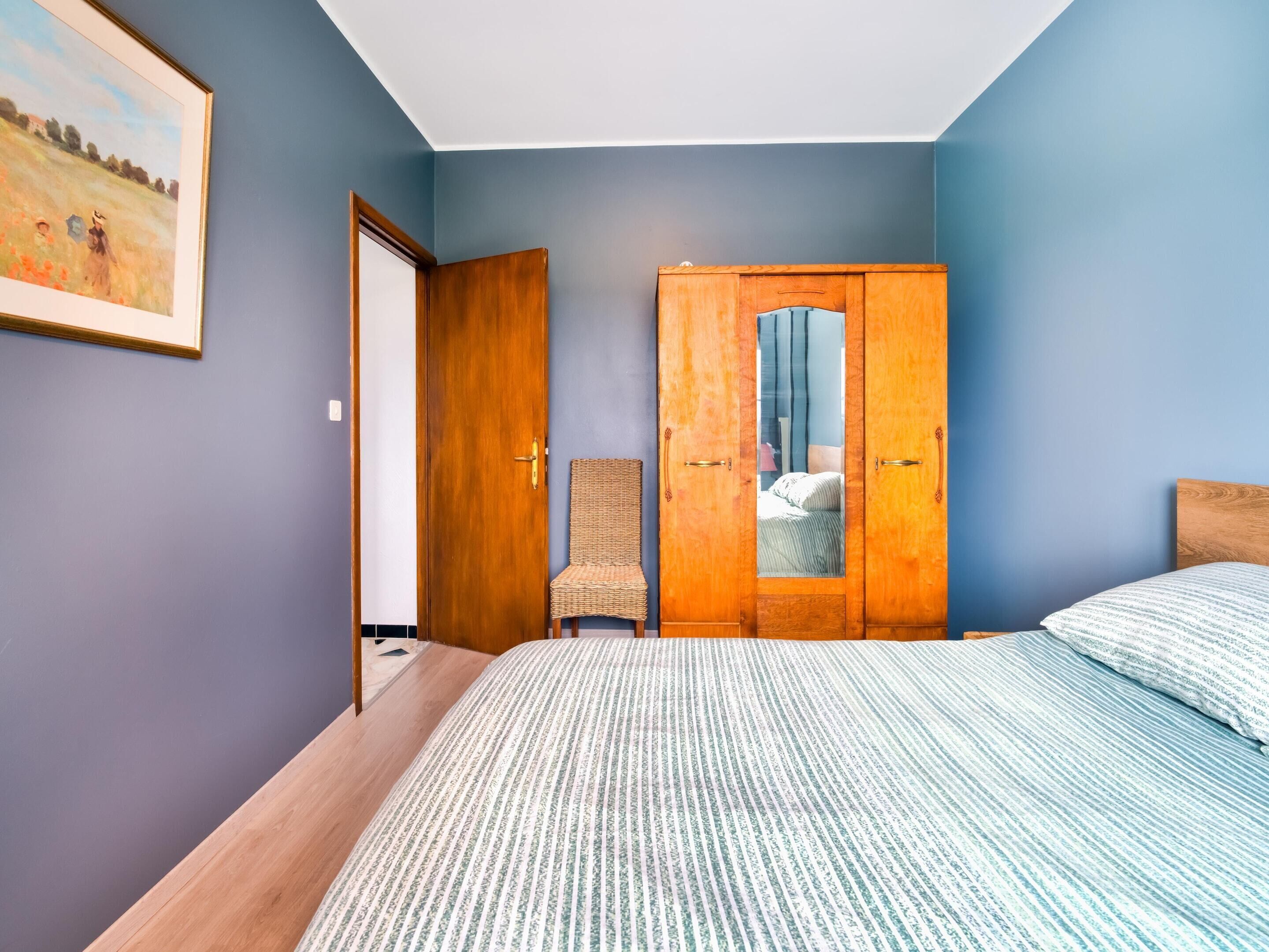 Une chambre à coucher avec un lit, une armoire en bois, une chaise en osier et un tableau accroché au mur.