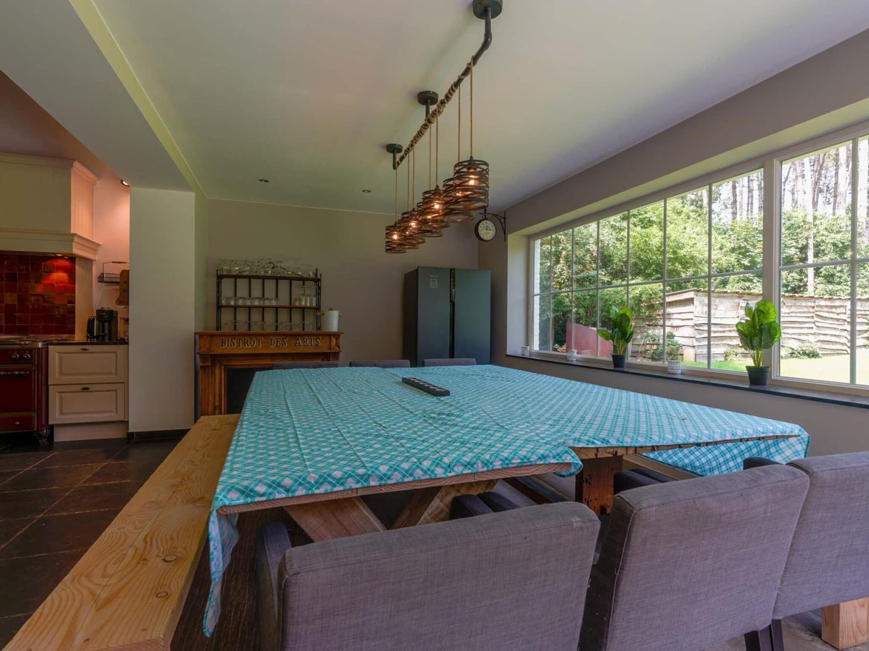A dining area with a table set for four, a kitchen with a stove, and a view of the outdoors through large windows.