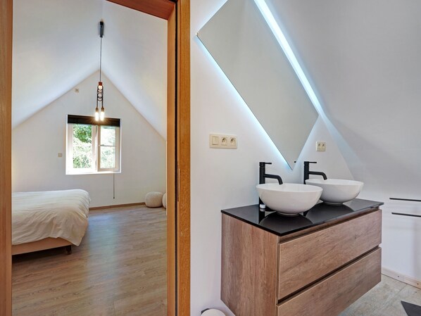 A modern bedroom with a sloped ceiling, a bed, a wooden dresser with a mirror, and a window with a view of greenery.