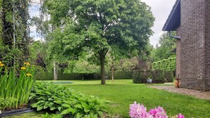 Un jardin bien entretenu, agrémenté de diverses plantes et fleurs, d’une pelouse et d’un grand arbre qui offre de l’ombre.