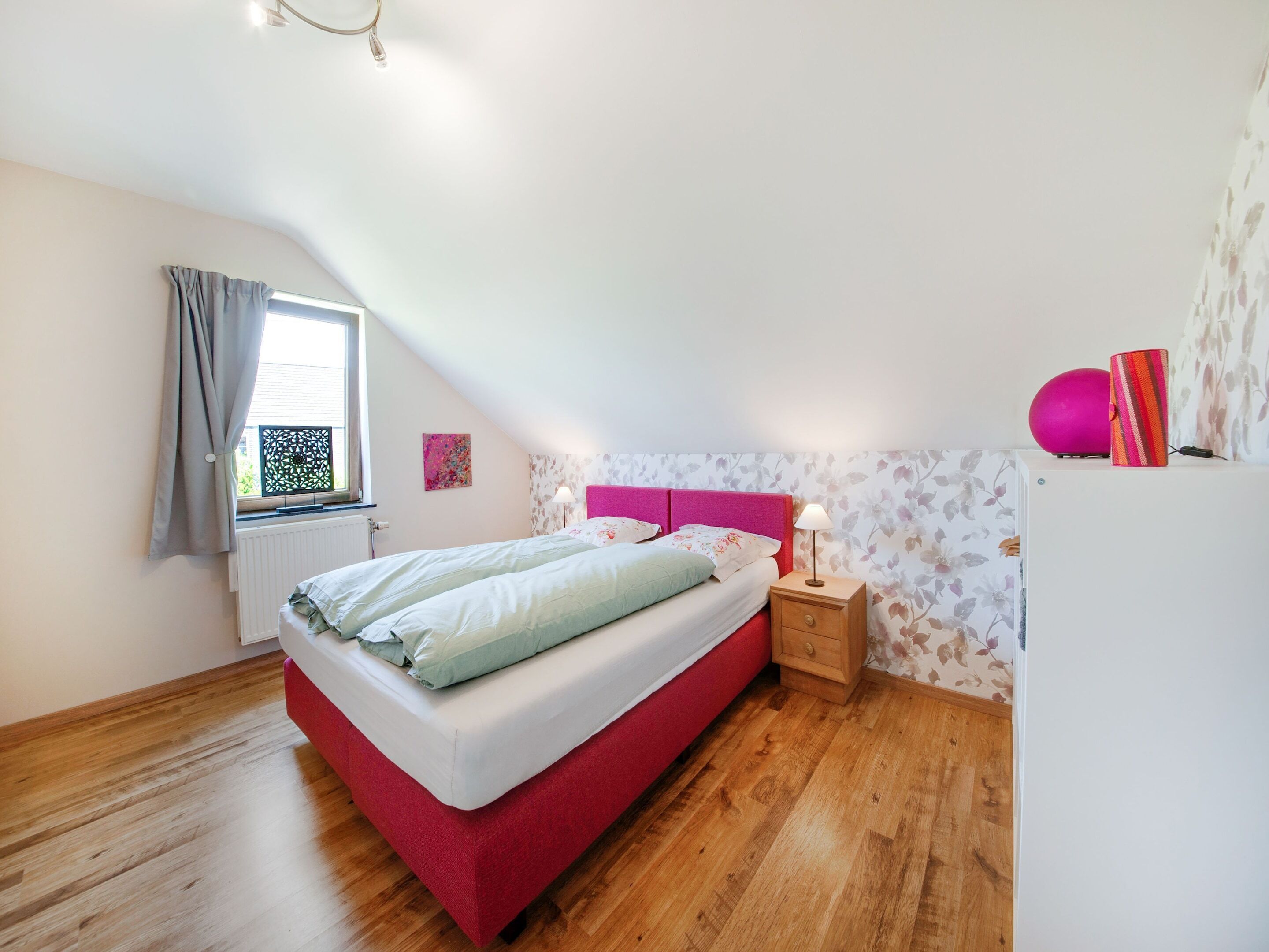 A bedroom with a red bed, a nightstand with a lamp, a window with curtains, and a radiator.
