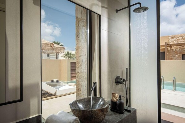 A modern bathroom with a large window, a stone basin, a shower, and a view of a pool and outdoor area. - Socrates Hotel (Hersonissos)